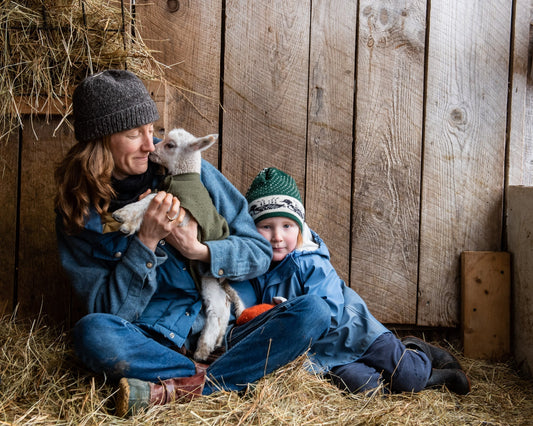 A woman holds a tiny sheep while a toddler leans on her